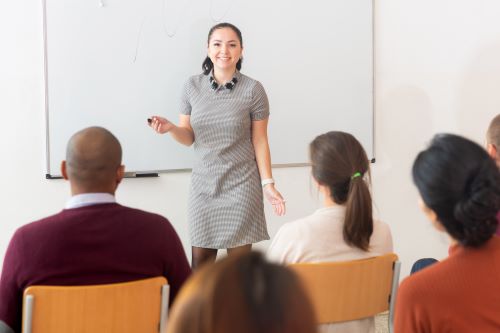 Frau unterrichtet vor einer Tafel und spricht zu den sitzenden Schulern deutsche Worter im Englischen skrivanek