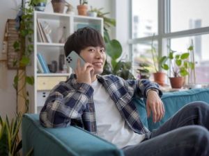 Ein japanischer Mann sitzt entspannt auf dem Sofa, lachelt und telefoniert mit seinem Smartphone hallo auf japanisch skrivanek