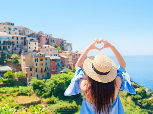 Eine Frau mit Strohhut formt ein Herz mit den Handen, Kustendorf und Meer im Hintergrund danke auf italienisch ubersetzungsburo berlin