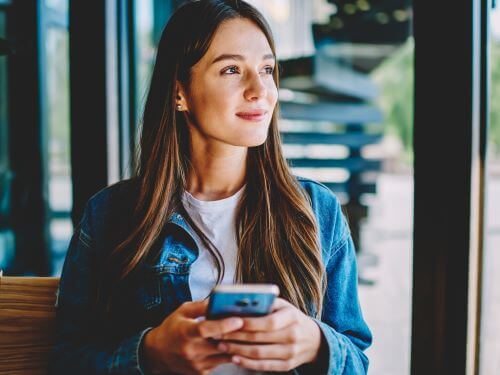 Junge Frau mit Smartphone schaut nachdenklich aus dem Fenster smiley danke ubersetzungsburo berlin