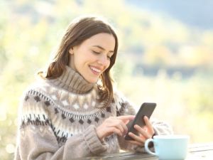 Lachelnde Frau in warmem Pullover nutzt ihr Smartphone bei einer Tasse Kaffee in einer naturlichen Umgebung was ist ecosia ubersetzungsburo berlin